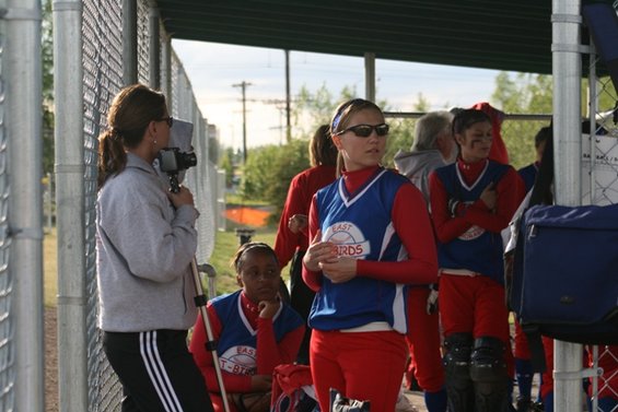 Softball players in red and blue uniforms stand near a fence