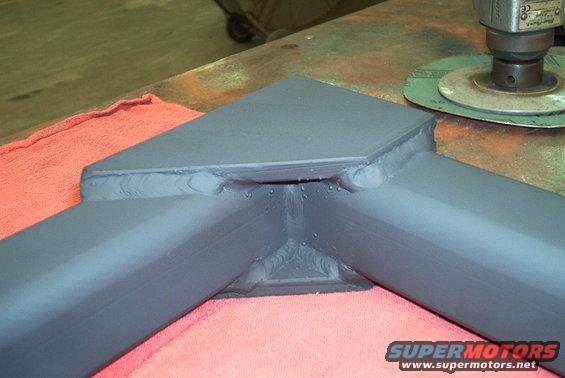 Close-up of a gray, welded metal frame with a buffing tool in the background