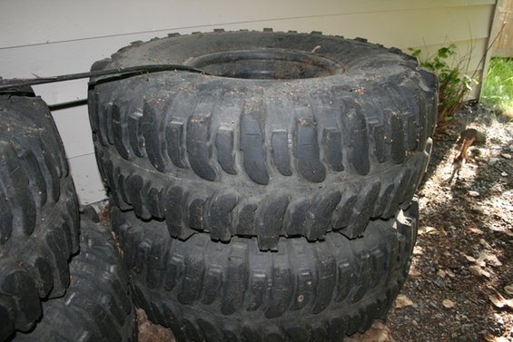 Stack of three large, knobby off-road tires, dusty and worn, leaning against a white wall