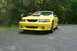 2002 Ford Mustang - A yellow 2002 Ford Mustang is parked on an asphalt surface with trees in the background.