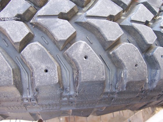 Close-up of aggressive off-road tire tread with deep lugs and siping. Small holes are visible in some lugs