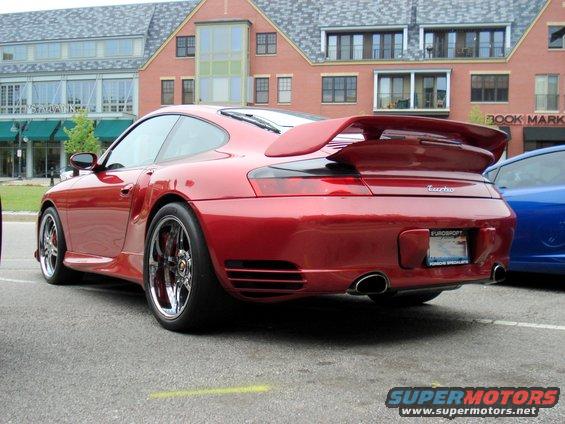 Red Porsche 911 Turbo with a large rear spoiler, seen from the rear three-quarter angle. A blue car is parked to its right