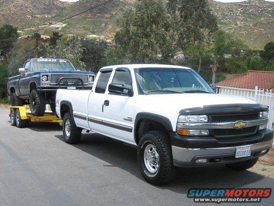 White pickup truck towing a blue pickup truck on a trailer. The white truck has a black bug deflector and chrome grille