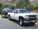 1979 Chevrolet C/K Pickup - A white pickup truck tows a blue pickup truck on a trailer down a paved road with hills in the background.