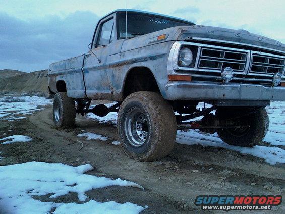Lifted pickup truck on a dirt road with snow patches. Front end shows a chrome grille and round headlights