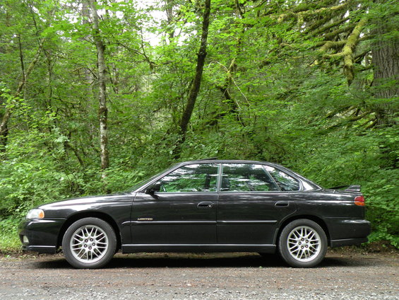 Black 1998 Subaru Legacy sedan, side view, parked on a gravel road with trees in the background
