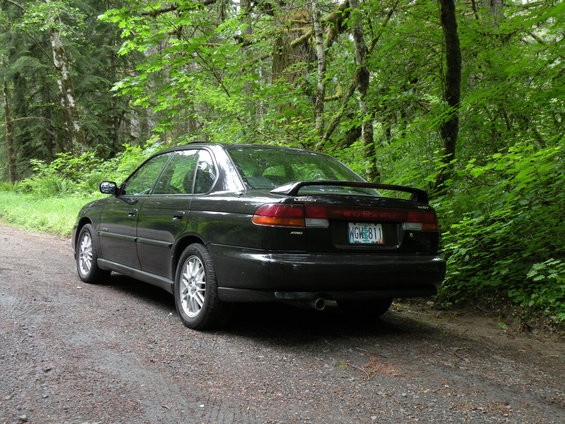 Black Subaru Legacy sedan parked on a dirt road in a forest. License plate reads KGH 811