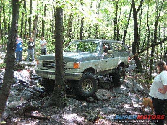 Two-tone gray and blue Ford Bronco navigating a rocky trail, with people watching