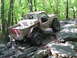 1991 Ford Bronco - An off-road vehicle with large tires is shown navigating a challenging rocky trail through a wooded area.