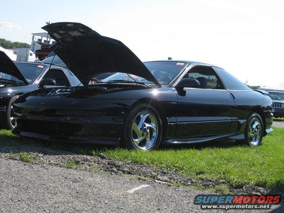 Black coupe with hood open, chrome wheels, parked on grass next to another car