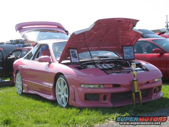 Pink 1994 Ford Probe with custom body kit, white wheels, hood and trunk open, trophy on display