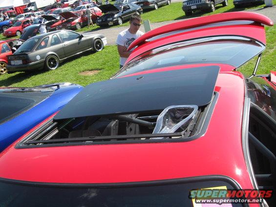 Red car with sunroof open, parked next to a blue car. Other vehicles and a person are visible in the background