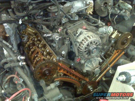 Engine bay with timing chains and alternator exposed. Supermotors.net logo visible