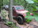 1991 Ford Bronco - A red Jeep Wrangler tackles a challenging muddy off-road course, followed by other vehicles.