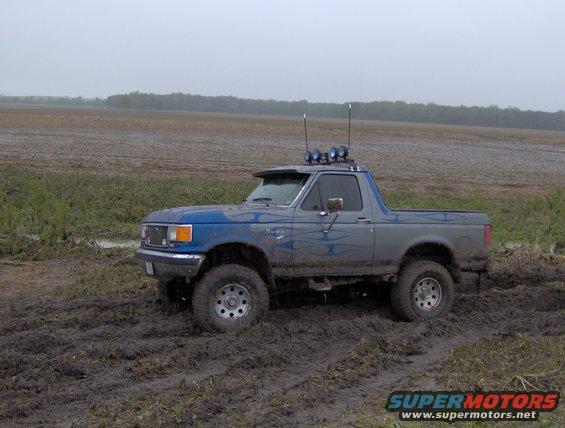 1987 Ford Bronco driving through a muddy field with aftermarket lights on the roof