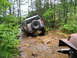 1991 Ford Bronco - A silver SUV tackles a challenging off-road course, splashing through a muddy stream surrounded by lush forest greenery.
