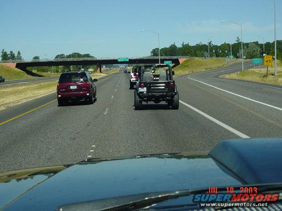 Rear view of a black Jeep Wrangler on a highway, following a maroon SUV. Another Jeep is visible ahead