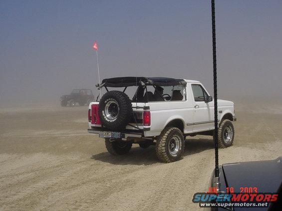 White Ford Bronco with spare tire on back, driving on a sandy beach with another vehicle in the distance