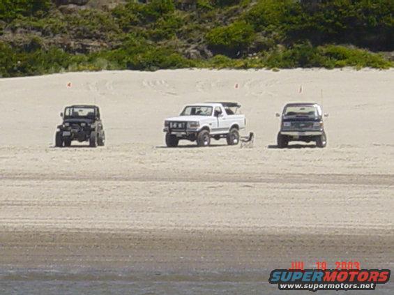 Black 1995 Jeep Wrangler, white Ford Bronco, and blue Ford Econoline van parked on a sandy beach