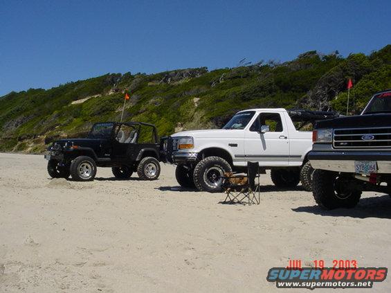 1995 Jeep Wrangler and two Ford Broncos parked on a beach with a grassy hill in the background