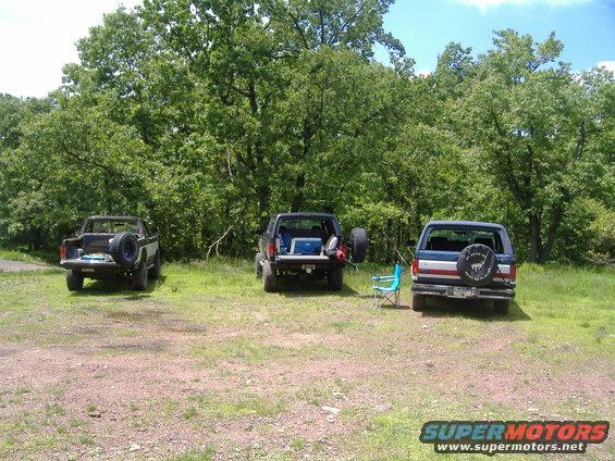 Three 1988 Ford Broncos parked in a grassy area with trees in the background. The middle Bronco has a cooler and camping gear in the open tailgate