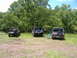 1988 Ford Bronco - Three 1988 Ford Broncos are parked in a grassy area with trees. The middle one has its tailgate open with gear inside.
