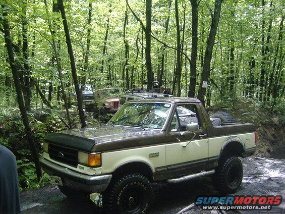 Two-tone brown and cream Ford Bronco with off-road tires, parked in a wooded area