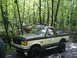 1988 Ford Bronco - A two-tone Ford Bronco navigates a wooded trail with other off-road vehicles in the background.