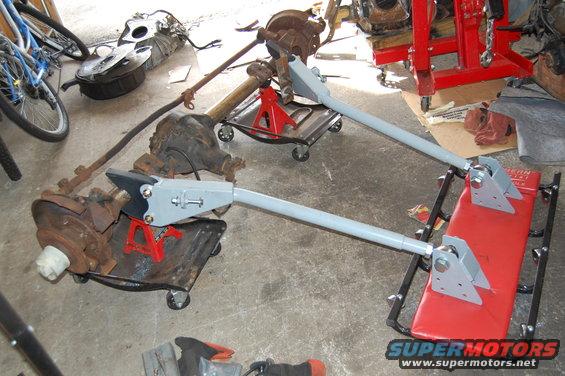 Two vehicle axle assemblies are supported by red jack stands and red creeper dollies, with a bicycle and other parts in the background