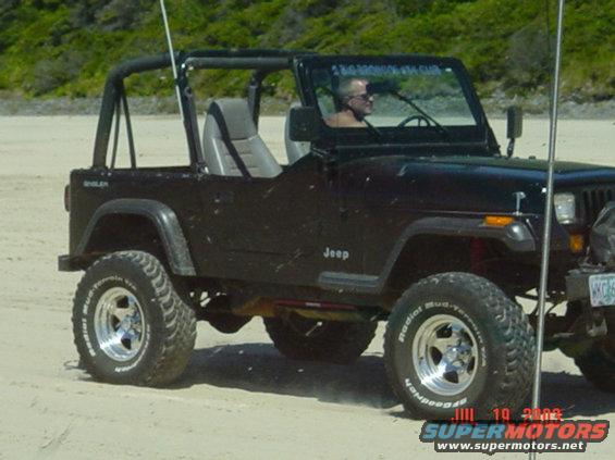 Black Jeep Wrangler on a sandy beach, driver visible, aftermarket wheels, soft top