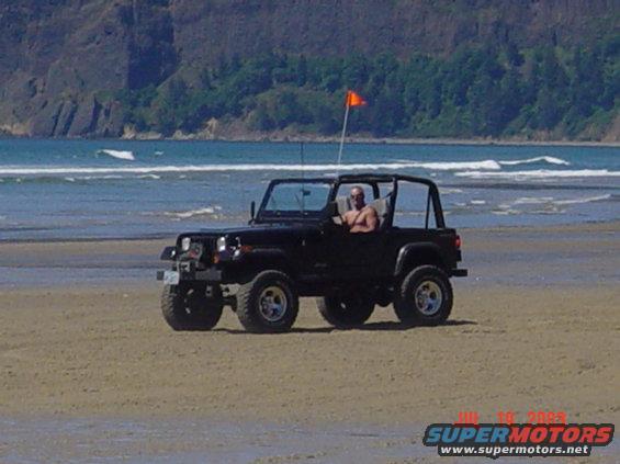 Black Jeep Wrangler with a man driving, parked on a sandy beach near the ocean. Orange flag on a pole attached to the Jeep