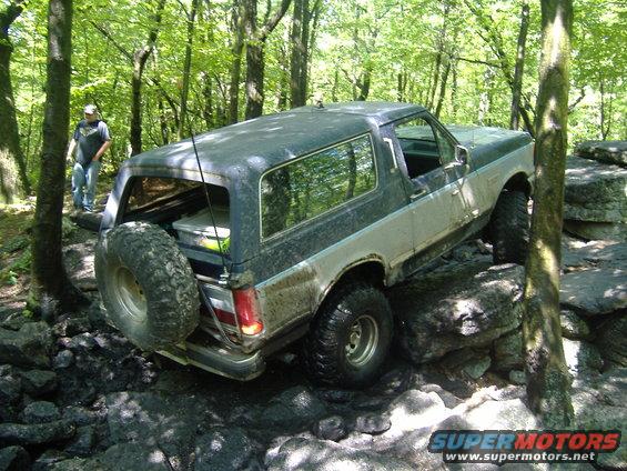 Ford Bronco climbing over rocks on a wooded trail. Spare tire mounted on the back