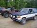 1988 Ford Bronco - A two-tone Ford Bronco is parked on a dirt road with a lifted pickup truck beside it.