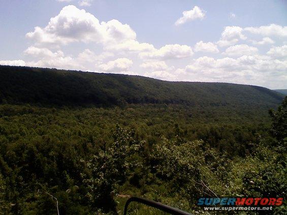 Forested hills and sky with clouds, a railing in the foreground. Watermark reads SUPERMOTORS.NET