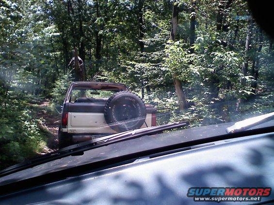 Brown and tan Ford Bronco with spare tire on rear, driving on dirt path in woods