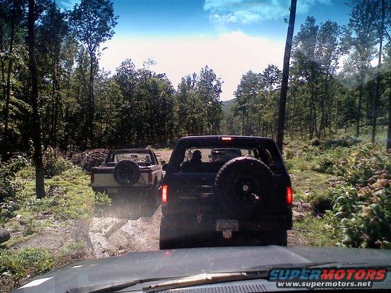 1988 Ford Bronco and a pickup truck driving on a muddy forest trail