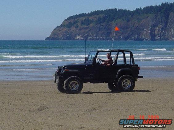 Black 1995 Jeep Wrangler driving on a beach with ocean and a hill in the background