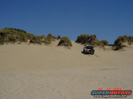 White SUV driving on sand dunes under a clear blue sky. Watermark reads JUL 10, 2003 SUPERMOTORS.NET