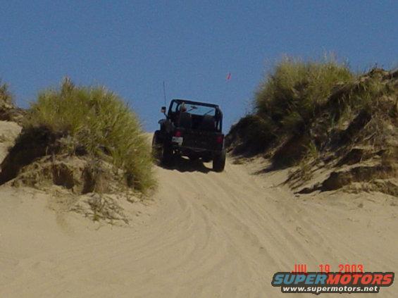 Black Jeep Wrangler driving up a sandy incline, rear view. The vehicle has a soft top and appears to be from the late 1990s