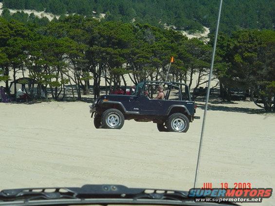 1995 Jeep Wrangler driving on a sandy beach with trees and dunes in the background