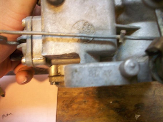 Close-up of a metal carburetor with visible stamped numbers and a hand holding a cable