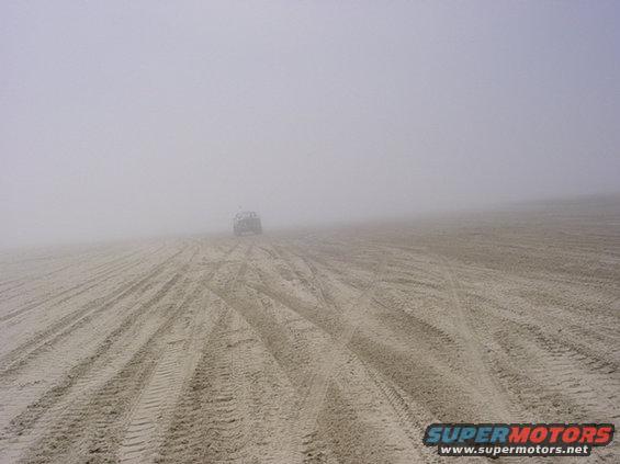 Vehicle driving on a sandy beach with tire tracks visible, obscured by fog