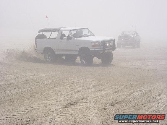 White 1995 Ford Bronco driving on sand with another vehicle behind it in fog