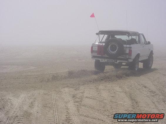 White Ford Bronco with spare tire on back driving on a sandy beach, red flag on antenna. Fog obscures the background