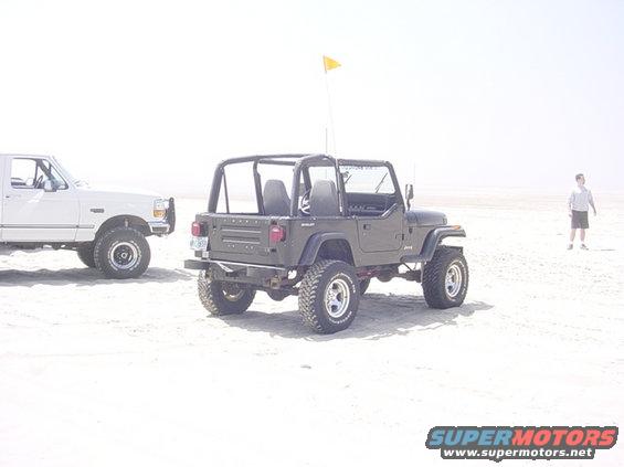 Dark green Jeep Wrangler, rear view, on a sandy beach with a white truck to its left