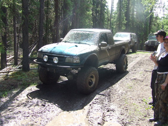 Muddy pickup truck with big tires on a dirt trail, other off-road vehicles and people in background