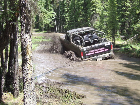 Muddy off-road vehicle with roll cage splashing through water, text on tailgate reads 'GOOD GIRL GONE REDNECK'