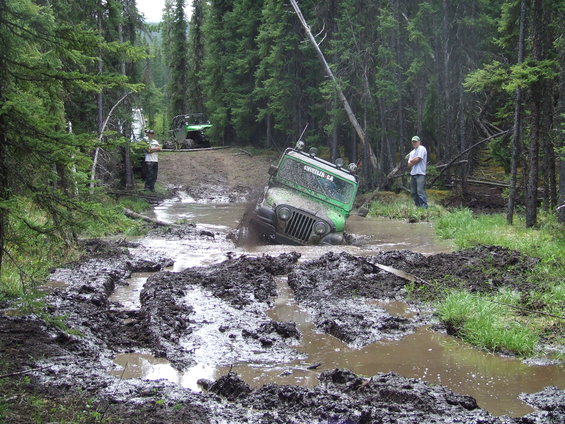 Green Jeep stuck in mud on a forest trail, with another vehicle in the background
