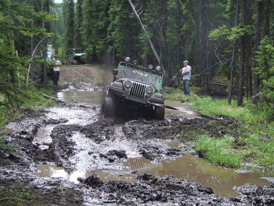 Green Jeep Wrangler struggling through deep mud on a forest trail. Another off-road vehicle is visible further up the path