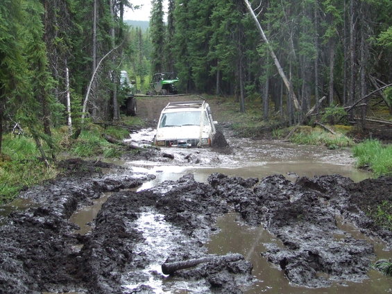 White SUV stuck in a muddy forest road. Two ATVs are visible further up the path